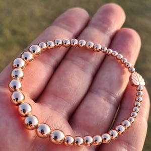 May include: A rose gold beaded bracelet with a small leaf charm. The bracelet is made of round beads and is stretchy.