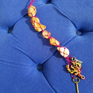 May include: A handmade beaded charm featuring a string of yellow stones encased in pink netting. The charm includes a cluster of colorful beads, a small metal key, and a decorative metal piece. The charm is displayed on a blue surface.