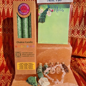 May include: A set of chakra candle, note pad, and crystal jewelry. The candle is green and has a label that says "Yogi & Yogini Naturals Chakra Candle". The note pad has a peacock design and says "Notes". The crystal jewelry is made of rose quartz and green aventurine.