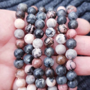 Puede incluir: Un conjunto de seis pulseras hechas con cuentas de piedras preciosas de color rosa, gris y negro. Las cuentas son redondas y tienen un patrón moteado.