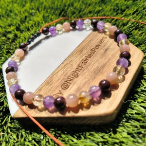 May include: A beaded bracelet featuring a variety of colorful gemstones, including purple amethyst, yellow citrine, and peach moonstone. The bracelet is displayed on a wooden and white surface with the text "@NightFairiesPowers".