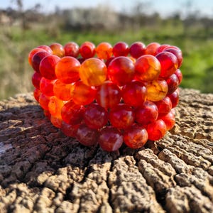 May include: A stack of red and orange beaded bracelets. The beads are round and appear translucent. The bracelets sit on a rough, textured wooden surface, with a blurred green background. The beads are approximately 1 cm in diameter.