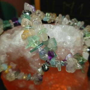 Op de afbeelding: Een veelkleurige kristallen armband met stukjes amethist, aquamarijn, fluoriet en helder kwarts. De armband wordt getoond op een roze Himalaya-zoutlamp.