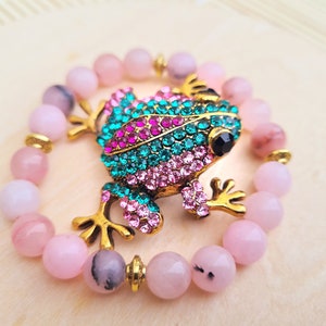 Könnte beinhalten: Ein Armband aus rosafarbenen Edelsteinen mit einem goldenen Frosch-Anhänger. Der Frosch ist mit rosafarbenen und grünen Strasssteinen verziert.