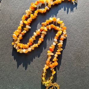 Può includere: Una collana d'oro con un ciondolo di pietra gialla e arancione. Il ciondolo è un simbolo del chakra del plesso solare. La collana è fatta di piccole pietre di forma irregolare.
