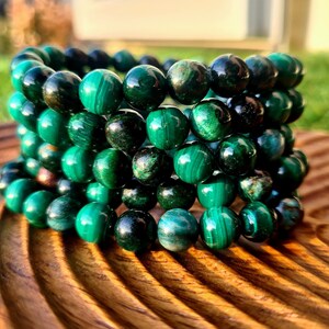May include: A stack of green malachite beaded bracelets. The beads are round and have a mix of dark and light green colors. The bracelets are displayed on a wooden surface.