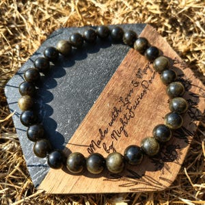 May include: A beaded bracelet featuring round, polished obsidian stones with golden sheen highlights. The beads are strung on an elastic cord, forming a circular shape. The text "Made with Love & Magic by Night Fairies Designs" is visible.