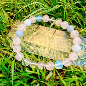 Pode incluir: Um bracelete feito de contas de pedras preciosas rosa, transparentes e azuis. O bracelete está sobre um leito de grama verde.