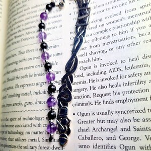 Puede incluir: Un marcapáginas de plata con un diseño trenzado y una cadena de cuentas negras y moradas. El marcapáginas tiene forma de serpiente y tiene un pequeño amuleto en el extremo. El marcapáginas está sobre un libro con texto sobre la deidad Yoruba Ogun.