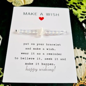 Pode incluir: Pulseira branca com um charme de estrela e um cartão que diz "MAKE A WISH" com um coração vermelho. O cartão também diz "put on your bracelet and make a wish. wear it as a reminder to believe it, seek it and make it happen. happy wishing!"