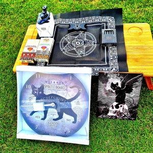 May include: A black and white spirit board clock with a black cat design. The clock has a black cat with a spirit board design on the face. The clock is in a white box. There is a black and white cat with wings on a skull on a black and white background. There is a black cloth with a white pentagram design. There are two boxes of incense sticks.