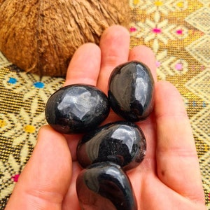 Peut inclure: Quatre pierres lisses et noires, probablement de l'obsidienne ou de la tourmaline noire, sont tenues dans une main. Les pierres sont polies et ont une finition brillante.