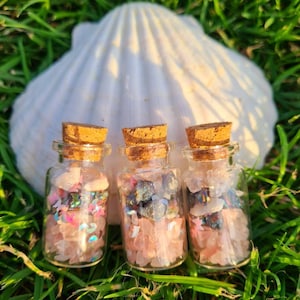 May include: Three small glass bottles with cork tops filled with pink, white, and blue crystals and glitter. The bottles are sitting on a bed of green grass with a white seashell behind them.