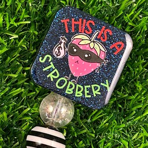 May include: A badge reel with a square, glittery navy blue top featuring a cartoon strawberry wearing a mask and holding a money bag. The text reads "THIS IS A STROBBERY" in red and green. The reel has decorative beads.