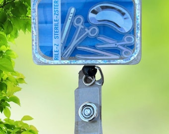 May include: A blue and silver badge reel with a clear plastic cover featuring a medical illustration of surgical instruments. The text "RULE-7 STERILE-STERILE" is printed on the badge reel.