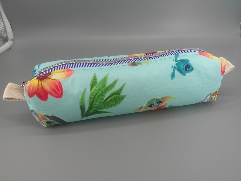 May include: A cylindrical pencil case with a light blue background, featuring a colorful floral and character pattern. The design includes red, orange, and yellow flowers, green plants, and cartoon characters. A zipper runs along the top, and a beige fabric loop is attached to the side.