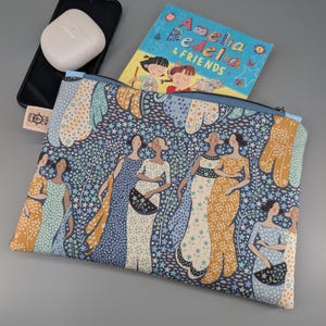 May include: A blue zippered pouch with a repeating pattern of women in dresses. The pouch contains a children's book titled "Amelia Bedelia & Friends." A smartphone and wireless earbuds are visible next to the pouch.