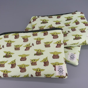 May include: Two zippered pouches with a light green background and a repeating pattern of a cartoon character in brown robes. The pouches have black zippers and a white tag with a logo. The pouches are in different sizes.