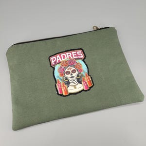 Könnte beinhalten: Eine olivgrüne Reißverschlusstasche mit einem zentralen gestickten Aufnäher. Der Aufnäher zeigt eine Figur mit Totenkopf-Make-up, einem Blumenkranz und Kerzen, wobei das Wort PADRES in weißen Buchstaben auf rosa Hintergrund darüber steht.