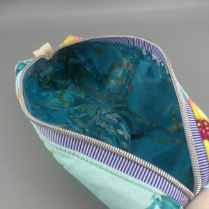 May include: Open cosmetic bag with a floral design. The exterior is light blue with a sunflower pattern on the corner. The interior is a vibrant blue with a marbled effect. A silver zipper and striped trim complete the design.