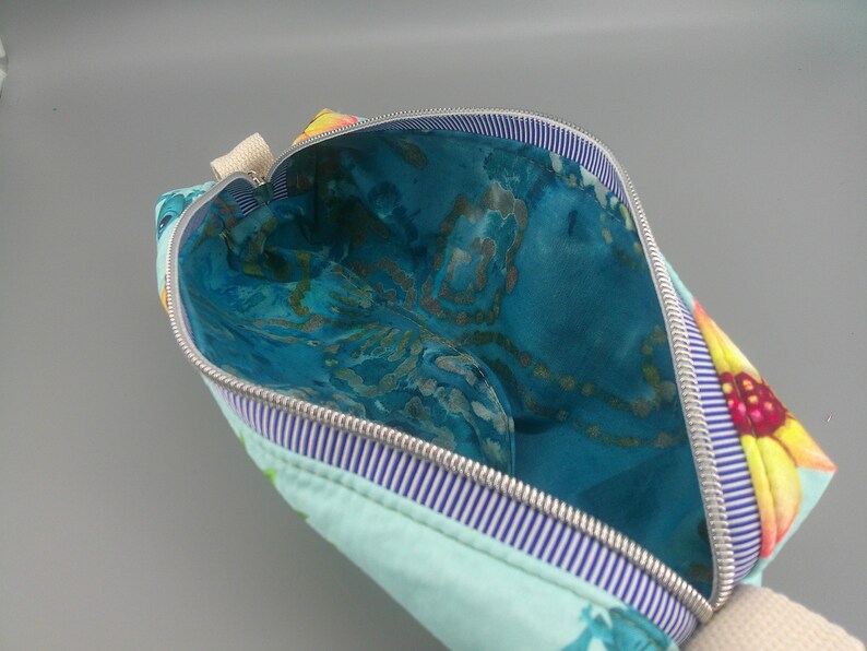 May include: Open cosmetic bag with a floral design. The exterior is light blue with a sunflower pattern on the corner. The interior is a vibrant blue with a marbled effect. A silver zipper and striped trim complete the design.