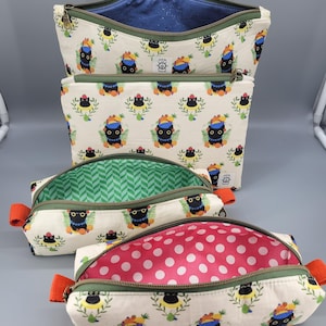 May include: A set of four zippered pouches in varying sizes featuring an off-white fabric with a repeating pattern of black cat heads adorned with colorful fruit. The pouches have olive green zippers, orange side tabs, and distinct interior linings.
