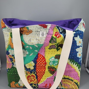 May include: A tote bag featuring a multicolored floral and geometric kantha quilt pattern with visible running stitch details. The bag has two wide cream-colored fabric straps and a solid purple interior lining. A small white label reads HEM AND HELM.