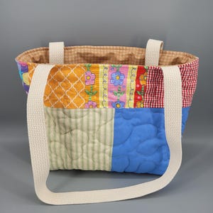 Puede incluir: Un bolso de tela acolchado con diseño de parches que presenta varios patrones coloridos, incluyendo celosía naranja, estampados florales, cuadros vichy rojos, rayas verdes y azul sólido. Tiene dos asas de tela tejida en color blanco roto.