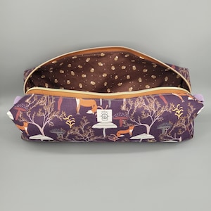 May include: A rectangular zippered pouch with a dark purple exterior featuring a print of orange foxes, trees, and mushrooms. A white square label with the text HEM AND HELM is centered on the front. The interior is lined with brown fabric.