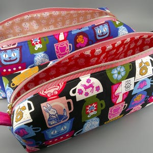 Puede incluir: Dos estuches con cremallera y estampados coloridos. Uno tiene fondo negro con tazas apiladas e interior rojo. El otro tiene fondo azul con tazas e interior beige. Ambos tienen ribete a rayas rojas y blancas.