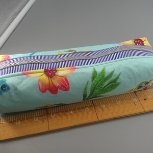 May include: A rectangular pencil case with a floral and sea creature pattern in shades of teal, yellow, and red. The case has a blue and white striped zipper and a beige fabric handle. A wooden ruler is next to the case.