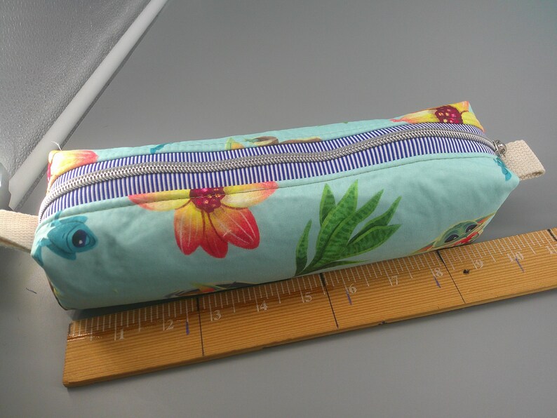 May include: A rectangular pencil case with a floral and sea creature pattern in shades of teal, yellow, and red. The case has a blue and white striped zipper and a beige fabric handle. A wooden ruler is next to the case.