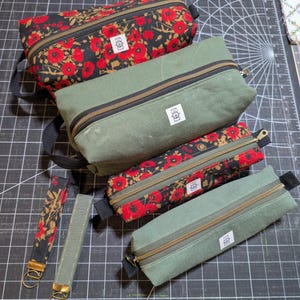 May include: Two rectangular zippered pouches with black fabric straps and two matching wristlet key fobs. One pouch and one fob feature a black floral pattern with red poppies. The other pouch and fob are solid olive green. Each item has a white label with the text Hem & Helm.