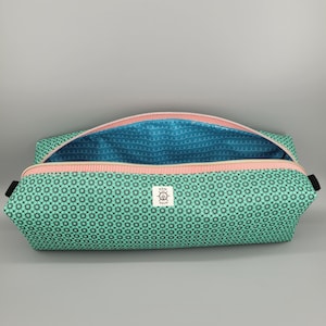 May include: A rectangular zippered pouch featuring a mint green exterior with a black geometric star pattern and a white label with the text HEM AND HELM. The interior is lined with bright blue fabric featuring a white swirling line pattern.