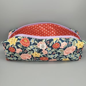 May include: A rectangular zippered pouch featuring a dark navy blue exterior with a multi-colored floral print of red, pink, and yellow flowers. The interior is lined with a coral fabric featuring a small white pattern. A label reads HEM AND HELM.
