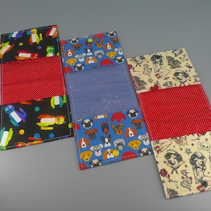 Puede incluir: Tres carteras de tela con diferentes estampados. Una tiene un fondo negro con personajes de dibujos animados coloridos, otra tiene un fondo azul con caras de perros y la tercera tiene un fondo beige con ilustraciones de estilo tatuaje. Cada cartera tiene un centro rojo con lunares.