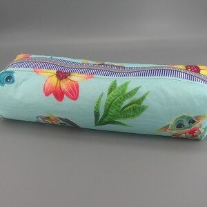 May include: A rectangular pencil case with a light blue background, featuring a floral and character print. The design includes large orange and red flowers, green leaves, and a cartoon character. The case has a striped zipper and a beige fabric handle.