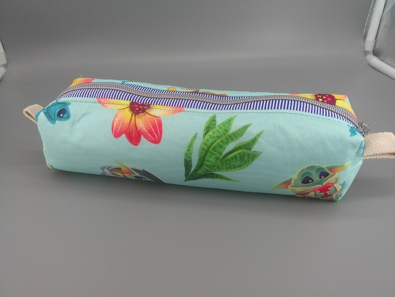 May include: A rectangular pencil case with a light blue background, featuring a floral and character print. The design includes large orange and red flowers, green leaves, and a cartoon character. The case has a striped zipper and a beige fabric handle.