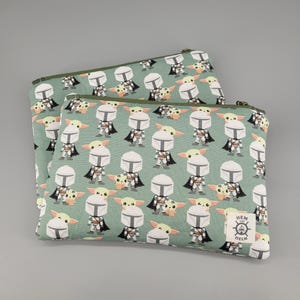 May include: Two rectangular zippered pouches in a light sage green fabric featuring a repeating pattern of cartoon characters in silver armor and small green creatures. A white rectangular label with the text Hem and Helm is on the front.