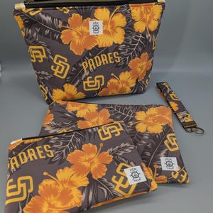 May include: A collection of Padres-themed accessories, including a large zippered pouch, two smaller zippered pouches, and a wristlet. The items feature a grey background with orange hibiscus flowers and the Padres logo. The accessories are made of fabric.