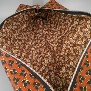 May include: Open orange tote bag with a black zipper. The exterior features a repeating butterfly pattern. The interior lining is a floral pattern with white and yellow flowers on a brown background. The bag is open, revealing the interior.
