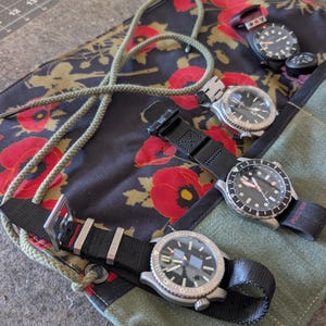 May include: A fabric watch roll featuring a black background with a red poppy floral pattern, secured by a green cord. The roll contains three wristwatches with black dials, silver-tone cases, and black nylon straps, alongside a small compass.