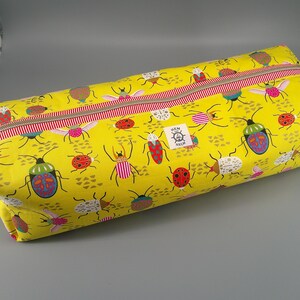 May include: A yellow rectangular zippered pouch with a repeating pattern of colorful insects. The insects are in various colors and designs, including ladybugs, beetles, and other bugs. The zipper is red and white striped.