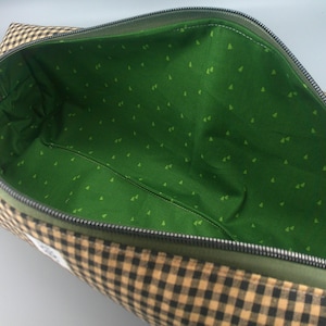 May include: Open view of a rectangular bag with a black zipper. The exterior is a brown and tan checkered pattern. The interior is green with small, light green triangle shapes. The zipper is black.