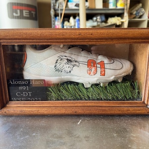 May include: A white football cleat with an orange number 91 and a black eagle design is displayed in a wooden shadow box. A black plaque with the text "Alonso Haro #91 C-DT WSE 2023" is visible below the cleat.