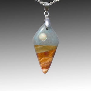 May include: A diamond-shaped pendant with a silver chain. The pendant features a light blue top section with a white pearl-like stone, and a layered, earthy-toned bottom. The chain is a silver, twisted rope style.