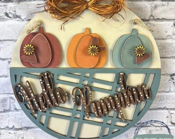 May include: A round wooden sign with three pumpkin cutouts in shades of red, orange, and teal. Each pumpkin has a sunflower and heart accent. The words "Hello Pumpkin" are cut out in a script font. A raffia bow and twine are at the top.