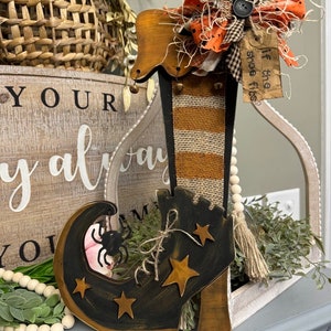 May include: A wooden Halloween decoration shaped like a witch's boot. The boot is black with gold stars and a spider accent. The boot has a burlap and orange striped shaft, and is topped with a bow and a tag that reads "If the shoe fits."