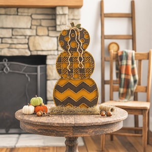 May include: A stacked pumpkin decoration featuring three tiers with distinct patterns: a top tier with polka dots, a middle tier with a plaid design, and a bottom tier with a chevron pattern, all in shades of orange, brown, and yellow.