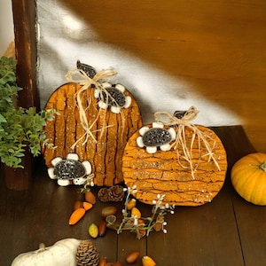 May include: Two wooden pumpkin decorations with textured orange surfaces and black and white accents. Each pumpkin has a raffia bow and decorative flowers. The pumpkins are on a dark wooden surface with other fall-themed decorations.